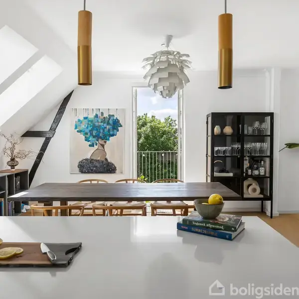 Et moderne køkken-alrum med et spisebord og stole. Der er en køkkenø med en skærebræt, citron og krydderurter. Baggrunden viser kunst, reol, planter og et åbent vindue med udsigt til træer.