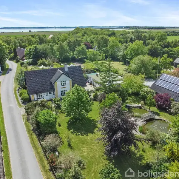 Hus omgivet af frodig have med dam og bro, beliggende ved en landevej i landligt område. Flere træer og buske i nærheden, solcellepaneler på nærliggende bygning.