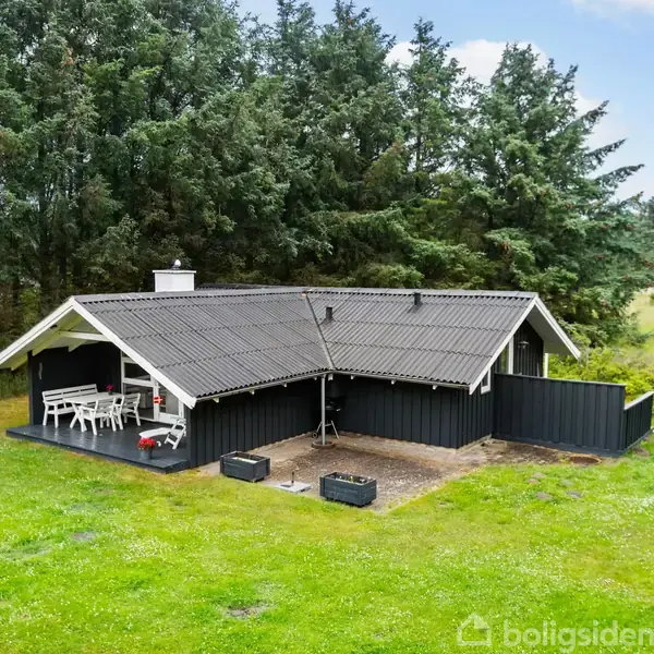 Sort sommerhus med gråt tag står stille på en grøn græsplæne omgivet af høje træer. En overdækket terrasse med hvide møbler ses ved siden af huset.