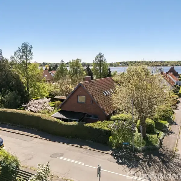 Hus med gavltag, omgivet af grønne træer og hække, beliggende i en rolig villavej nær en sø. Flere parceller og huse kan ses langs vejen under en klar blå himmel.