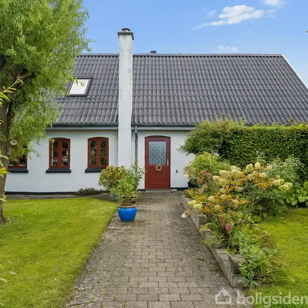 En hvid garage med en lukket port står ved siden af en brolagt indkørsel omkranset af grønne hække. I baggrunden ses et gråt hus under en blå himmel med få skyer.