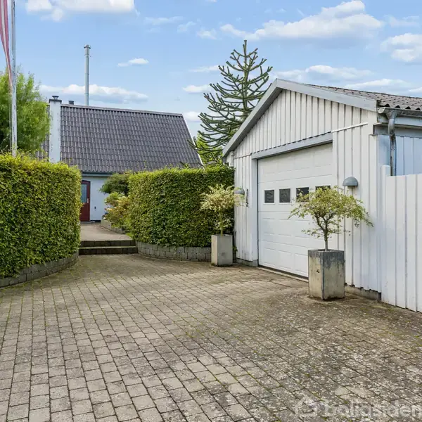 Et hvidt hus med mørkt tag står omgivet af velplejede grønne hække. Det ligger ved en vej i et grønt og frodigt boligområde.