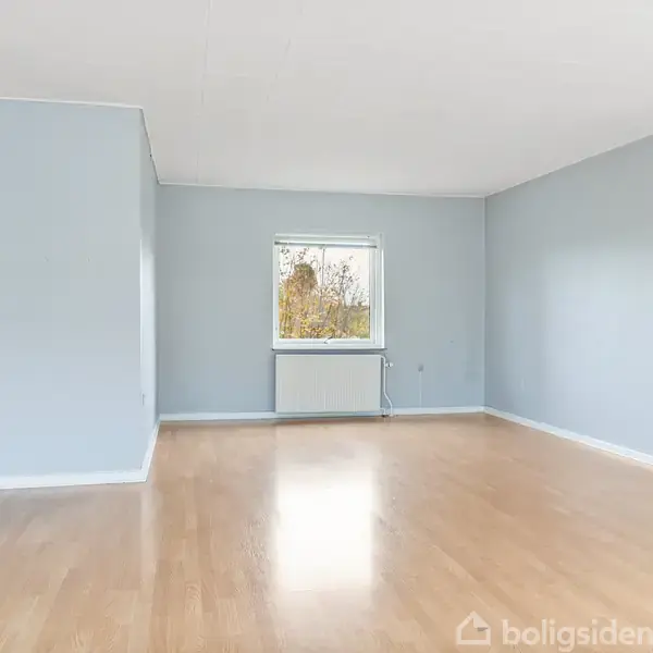 Et tomt rum med lyseblå vægge og lyst trægulv. Centralt er et vindue, der viser nogle træer udenfor. Rummet er minimalistisk uden møbler eller dekorationer.