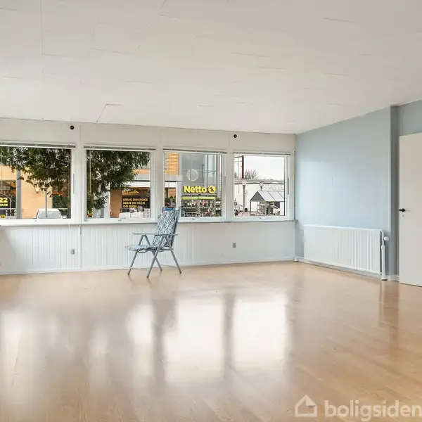 En enkelt stol står midt i et stort, lysfyldt rum med lyse trægulve og lyseblå vægge. Udenfor ses vinduer med udsigt til et Netto-skilt og træer.