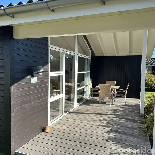 En træterrasse med fem havestole og et bord står under et halvtag, tilknyttet et sortmalet træhus. Nummer 35 ses ved indgangen. Sollys skinner på området.