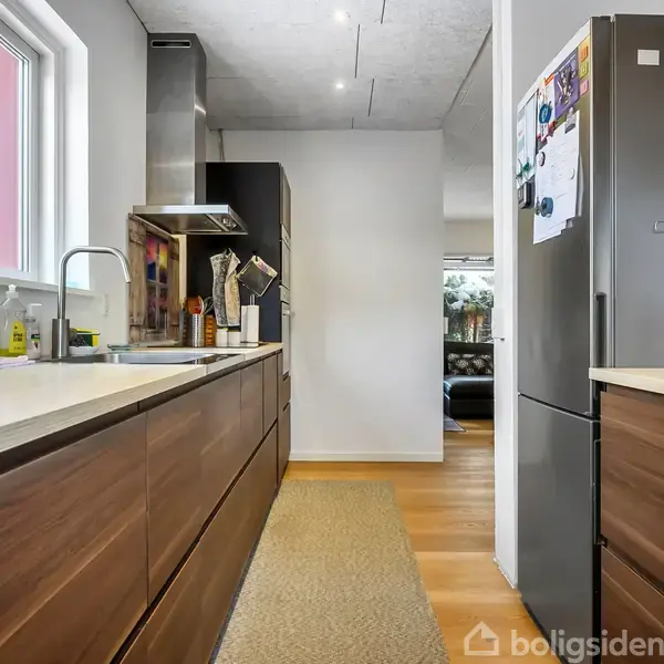 Brunt køkken med træskabe og stålhvidevarer, der står stille. En vask er til venstre, og et køleskab er til højre. Midt på gulvet ligger et tæppe.