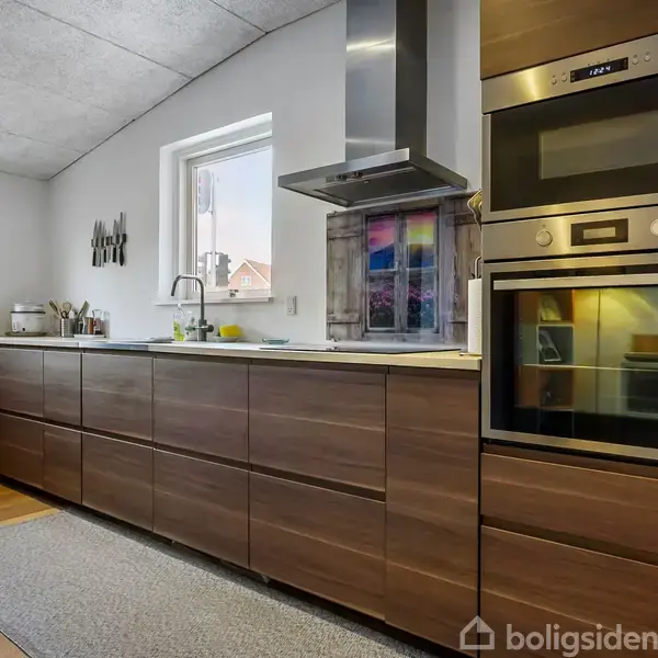 Moderne køkken med træskabe, indbygget ovn og mikrobølgeovn i en høj enhed. Et vindue ved vasken oplyser rummet, der har en enkelt loftlampe og en grå løber på gulvet.