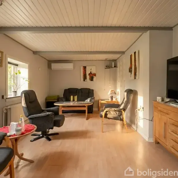 Stue med sofa og stole står omkring et træbord. TV på træskænk til højre, vindue til venstre og kunst på væggen. Duftlys og lamper giver hygge.