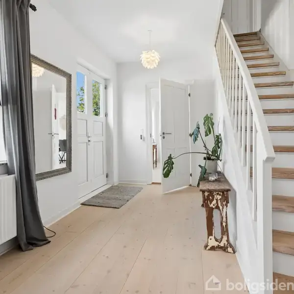 En lys entré med en trappe til højre. En plante står på et bord, og der ses et spejl og en lampe til venstre. Gangen fører til en åben dør.