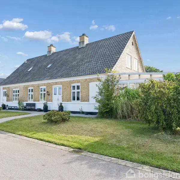 Et gult murstenshus med sort tegltag står stille på en velplejet græsplæne. En hæk omkranser huset langs vejen under en klar, skyfri himmel.