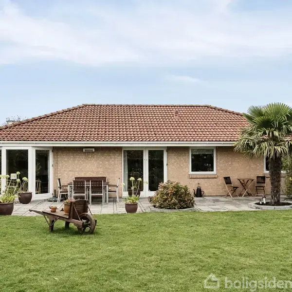 En villa med rødt tegltag står i centrum. Omgivet af en terrasse med havemøbler og krukker. Græsplæne og en palme i forgrunden. Himmel er klar.