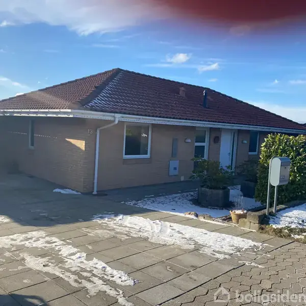 Et murstenshus med rødt tegltag står stille i en villavej. Der er en carport og en flisebelagt indkørsel, omgivet af lidt sne. Nummeret på huset er 13.