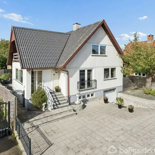 Hvid villa med sort tag står tom på en flisebelagt gårdsplads. Huset har trappeadgang og er omgivet af hegn. Et stort træ og en mindre have ses i baggrunden.