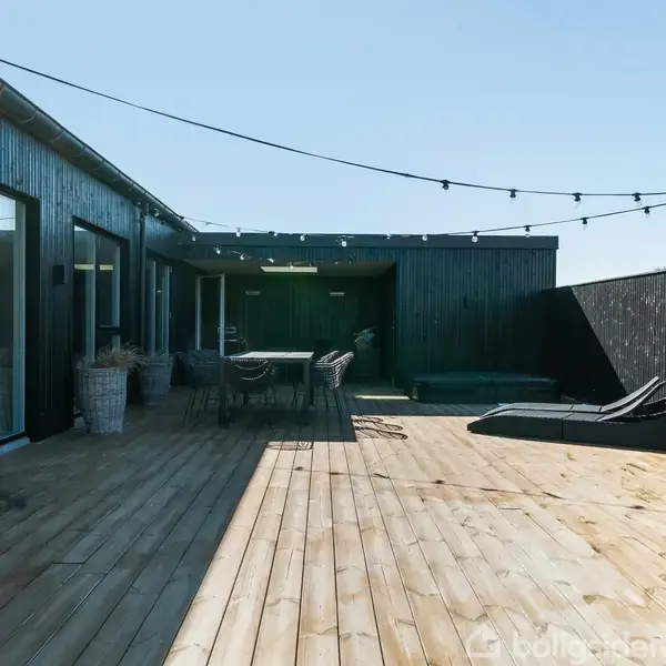 En stor træterrasse med møbler står foran et sort træhus. Lyskæder er spændt henover, og grønne planter ses i krukker. Det er solrigt og himlen er klar.