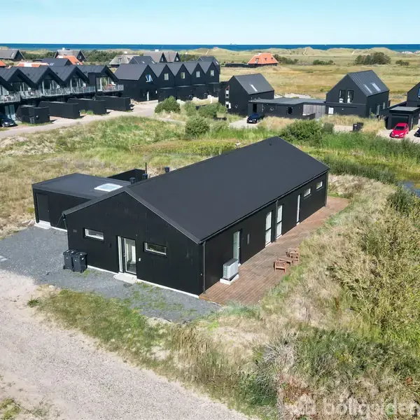 Sort, moderne hus står stille på en grusvej omgivet af græs og buske. I baggrunden ses flere lignende huse. Havet er synligt i horisonten.