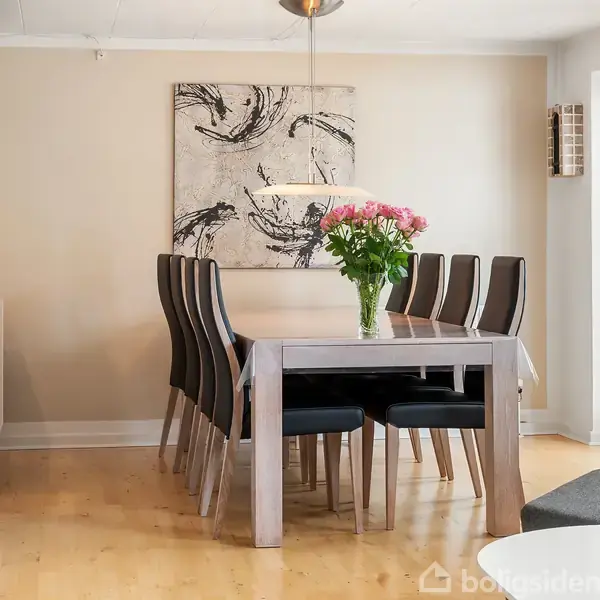 Et spisebord i træ er omgivet af otte stole, prydet med en vase med blomster. Væggene er dekoreret med et kunstværk, og der er en skænk til venstre.