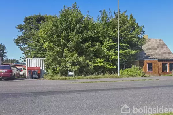 En vej løber foran et hus omgivet af træer og en container. To biler står parkeret ved siden af containeren i en landlig omgivelser.