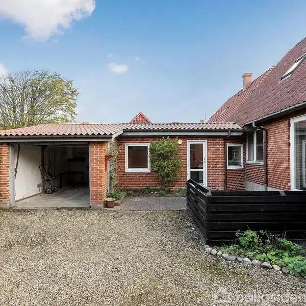 Rødt murstenshus i en gårdhave med en garage til venstre. Døren i midten har en plante ved siden af. Huset har rødt tegltag og en træterrasse til højre.