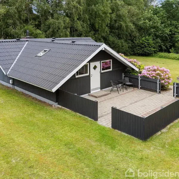 Sort træhus med skråt tag står på en flisesti indkapslet af et hegn. Der er en terrasse med havemøbler. Huset omgives af en græsplæne og træer i baggrunden.