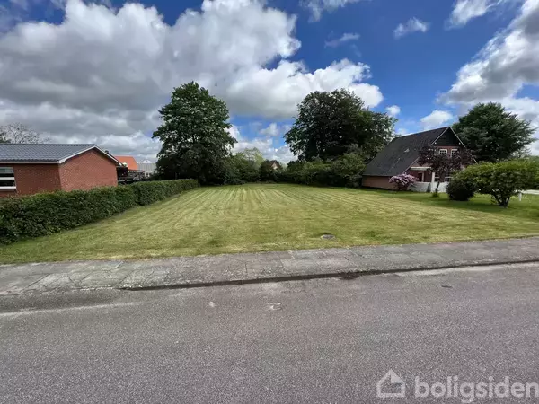 En græsplæne strækker sig foran to huse i et boligområde. Omkring plænen står store træer, og en vej løber i forgrunden. Skyer fylder himlen.