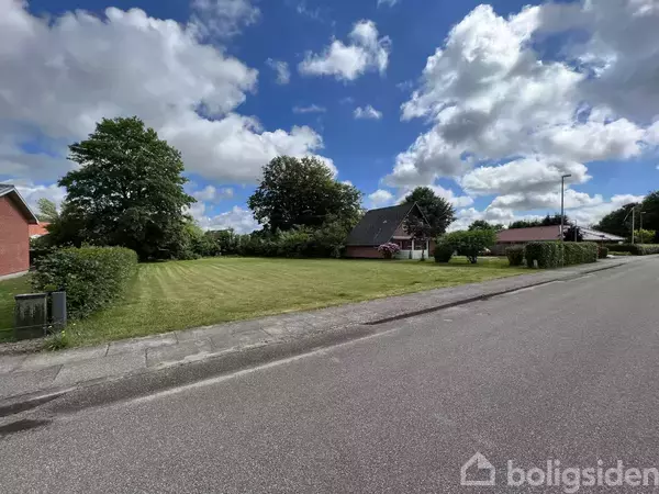 Velklippet græsplæne ligger stille ved siden af en vej, omgivet af buske og træer under en blå himmel med få skyer. I baggrunden ses et hus og flere træer.