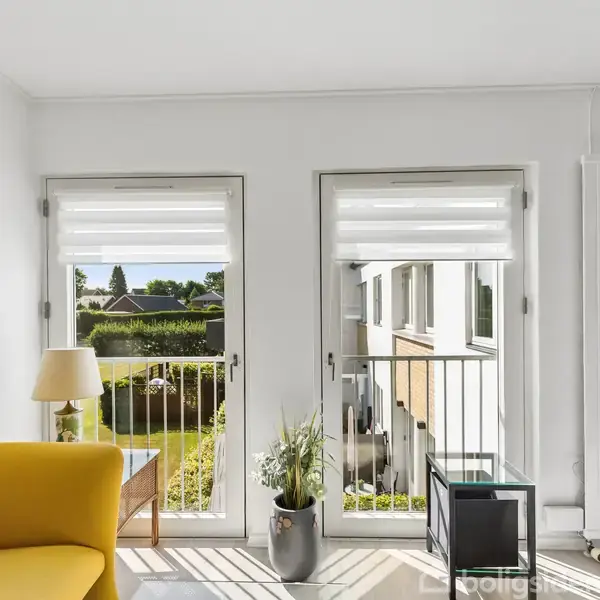 Gult sofaarrangement står i en lys stue med to vinduer, der åbner ud mod en altan. Udenfor ses en have med grønne buske. En plante og en lampe står ved vinduerne.