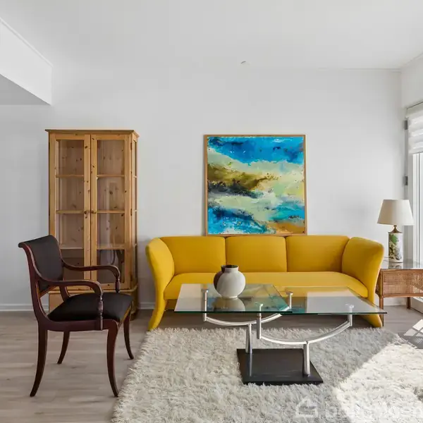 Gul sofa står i en stue foran et farverigt maleri. Glasbord med vase foran, omgivet af lænestole. Store vinduer lader naturligt lys strømme ind.