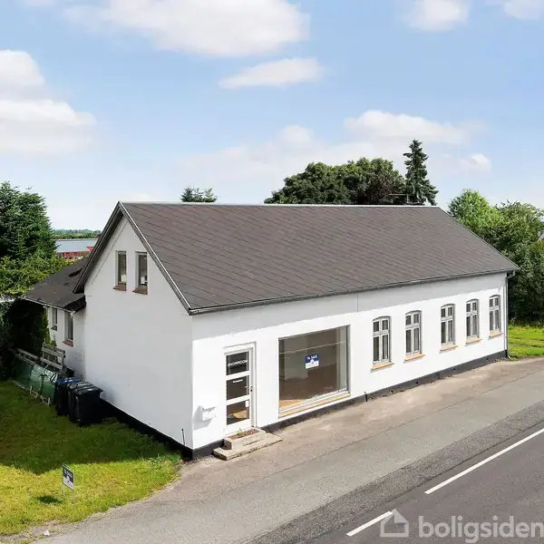 En hvid bygning med sort tag står ved en landevej. Den har store vinduer og en dør foran. Omgivet af grønne træer og en parkeringsplads med et vejskilt.