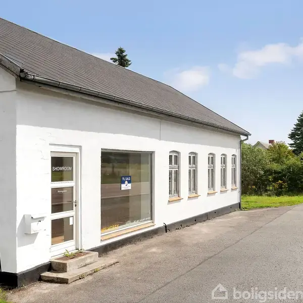 Bygning med hvid facade står stille ved en landevej. På døren står "SHOWROOM," og der er et "Til salg"-skilt i vinduet. Omkranset af asfalt og grønne træer.