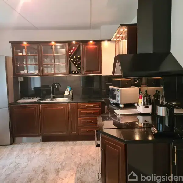 Køkken med mørke træskabe og sorte bordplader. En emhætte hænger over komfuret, mens en brødrister, mikroovn og kaffemaskine står på bordet. En metalfarvet køleskab ses i hjørnet.