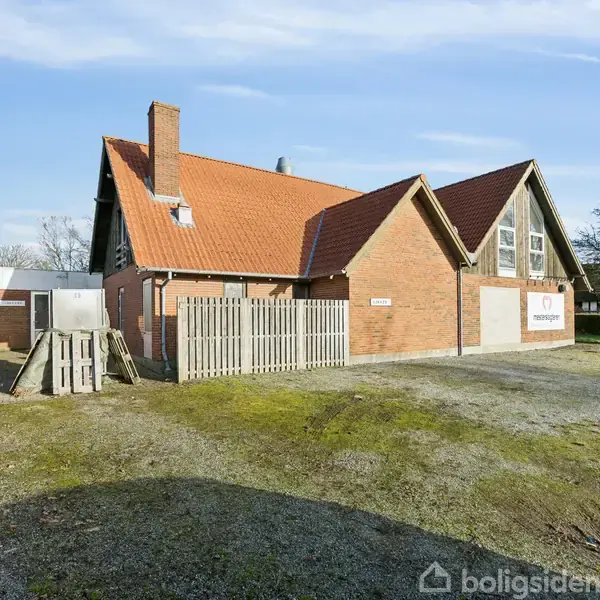 Rødt teglhus med skråt tag og skorsten står i fredelig gårdhave. Planter omkranser bygningen, mens et skilt på væggen viser teksten "Tømrerfirmaet".