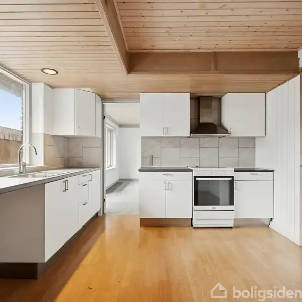 Et hvidt køkken med træloft og trægulv. Store vinduer lader naturligt lys ind. Der er et komfur med en emhætte, og hvide skabe langs væggene.