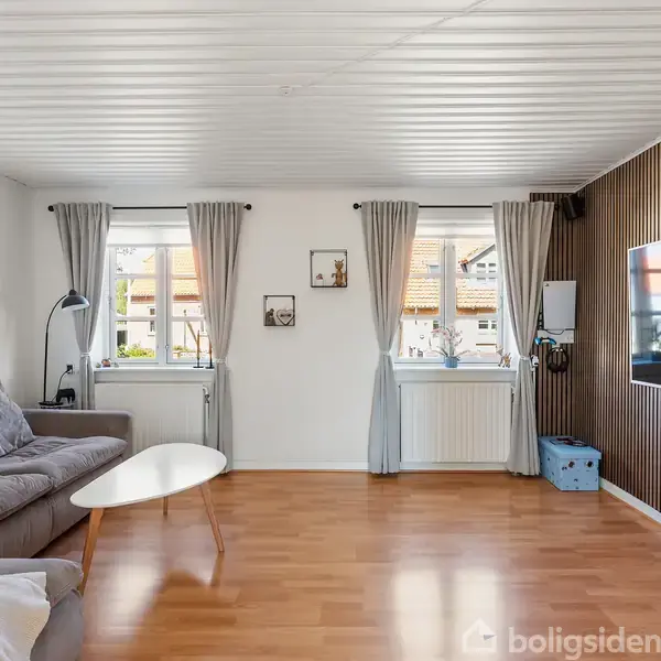 En stue med en grå sofa og et hvidt sofabord. En stor fladskærm hænger på en trælamelvæg. To vinduer trukket fra af grå gardiner slipper lys ind.