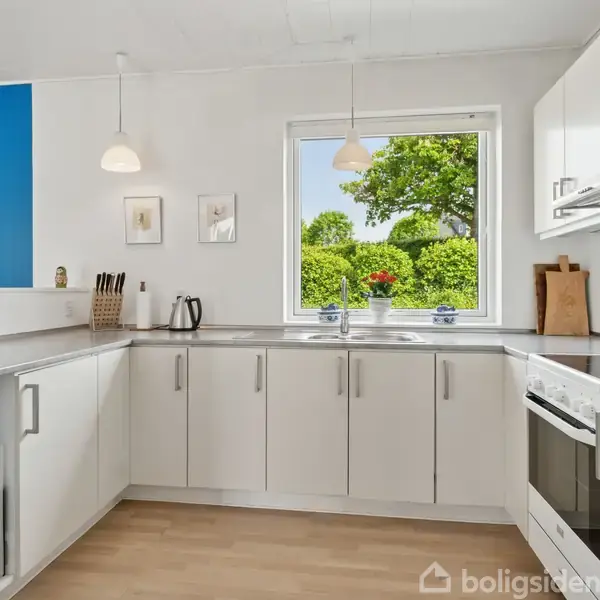 Rødt murstenshus med rødt tegltag står på stor grund. En garage med metaltag ved siden af huset. Grønne buske og træer omkring, landlig baggrund med marker og vej.