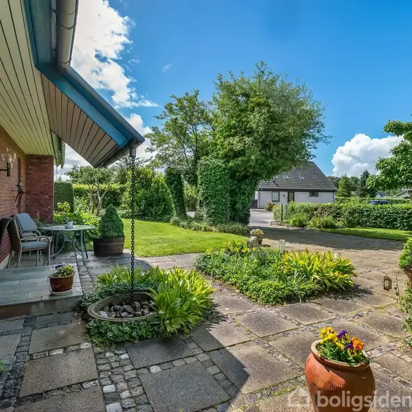 Hus med terrasse: Rød murstensvæg og terrassebord møbleret med stole og planter. Omgivet af frodig have, græsplæne, blomsterbede og anden bygning i baggrunden under en lys, blå himmel.