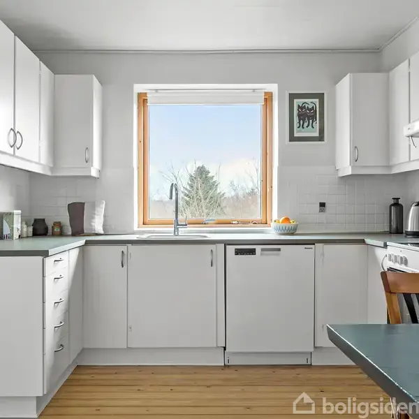 Et lyst køkken med hvide skabe og mørke bordplader. En kaffemaskine og brødrister står fremme. Ud over vinduet ses et træ og blå himmel.