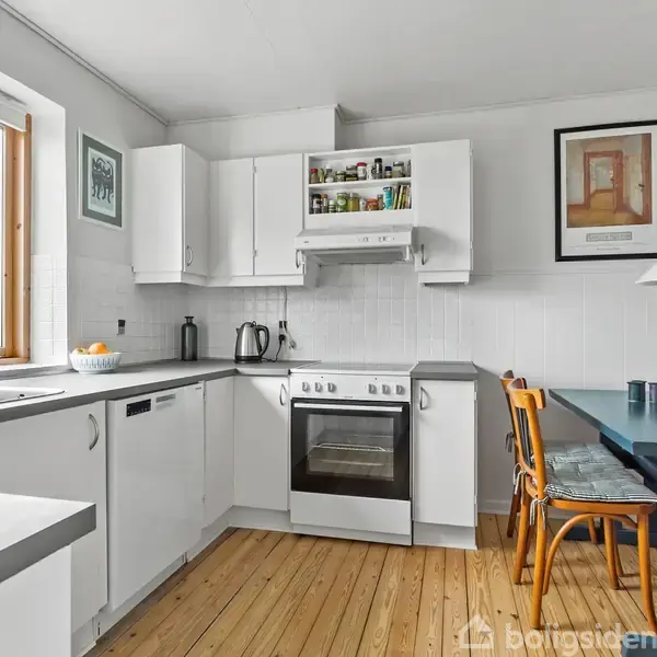 Et hvidt køkken med en komfur og ovn under hvide skabe. Ved siden af hænger et kunstværk på væggen. Et bord med træstole står ved vinduet, som har udsigt til træer.