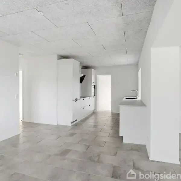Åbent, lyst køkkenalrum med hvide køkkenmoduler står i midten, gulvet har lyse fliser, og rummet er delvist opdelt af vægge. Hvidt loft med synlige linjer.