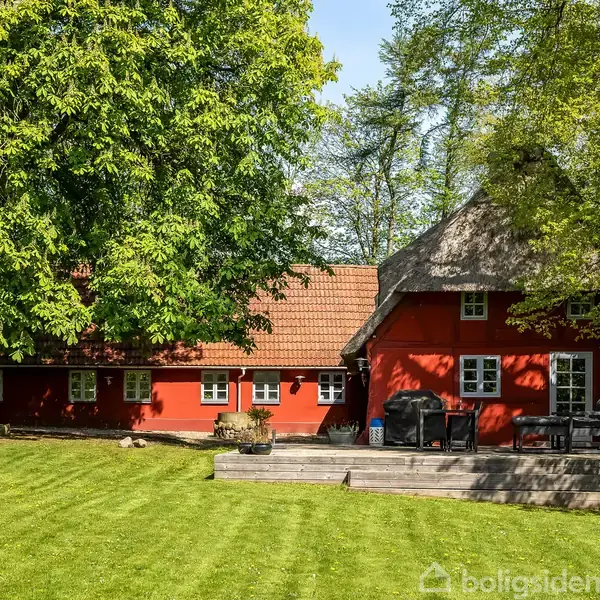 Et rødt bindingsværkshus står stille i en frodig, grøn have med tætte træer og en velplejet græsplæne. En terrasse foran huset har havemøbler til afslapning.