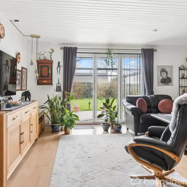 Stue med sort læderlænestol og sofa ved sofabord. TV og skænk på væggen. Store vinduer viser haven udenfor. Grønne planter dekorerer rummet med billeder og vægkunst.