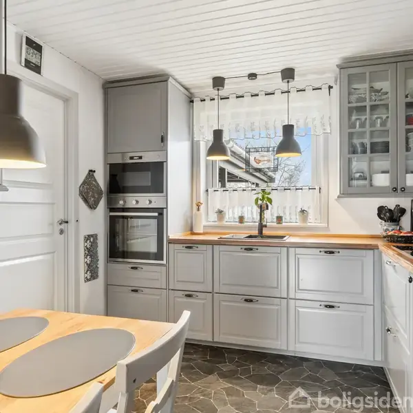 Køkken med lysegrå skabe og hvide fliser, indbygget ovn ved siden af et vindue med udsigt. På bordpladen står en plante. To pendellamper hænger over et lille spisebord.