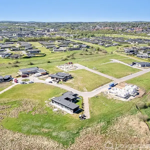 Luftfoto af et boligområde med flere moderne huse spredt ud over grønne græsarealer. Der er veje, og nogle huse er under konstruktion. Bylandskab ses i baggrunden under en klar blå himmel.