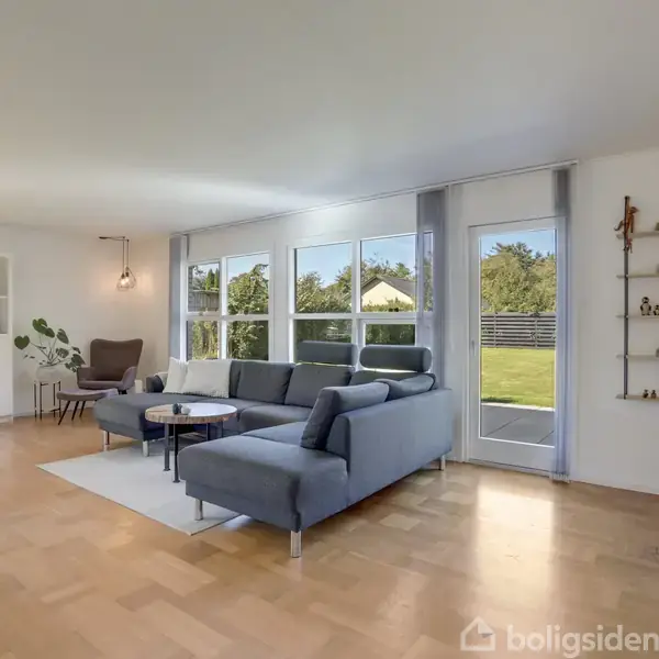 Sofa stående i moderne stue, badet i naturligt lys fra store vinduer med udsigt til en have. Rummet er indrettet med hvide skabe, en lænestol og åbne hylder.