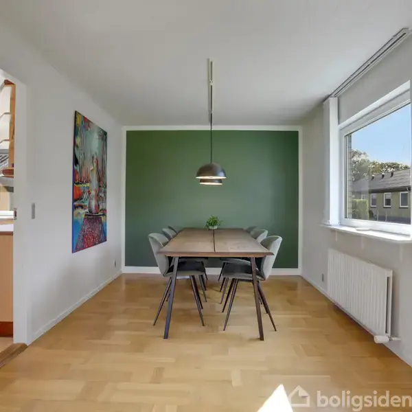 Et spisebord med seks stole står i et lyst rum med trægulv. En lampe hænger over bordet. Der er en grøn væg, et farverigt maleri, og et stort vindue.