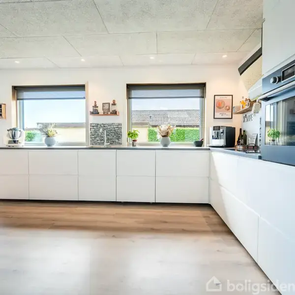 Et moderne køkken med hvide skabe og sorte vinduesrammer. Bordpladen er dekoreret med planter og køkkenapparater. I hjørnet ses to ovne. Detaljer skaber en lys og indbydende atmosfære.