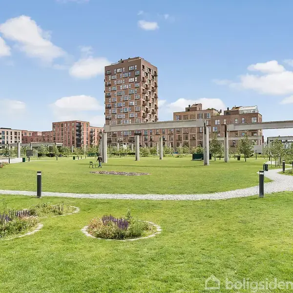 Grøn park med blomsterbede omgivet af moderne lejlighedsbygninger. Et betonpergola-struktur ses i midten, og stier krydser det åbne græsareal under en blå himmel.