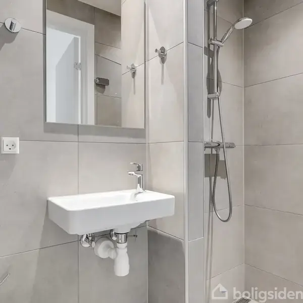 Håndvask hænger på væggen under et spejl i et moderne badeværelse med grå fliser. Brusehoved og kontrolpanel ses ved siden af, med en døråbning i baggrunden.
