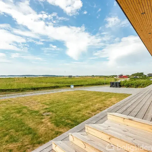 Træterrasse med trappe fører til en græsplæne, beliggende nær en kystlinje. I baggrunden ses åbent land og enkelte bygninger under en delvist skyet himmel.