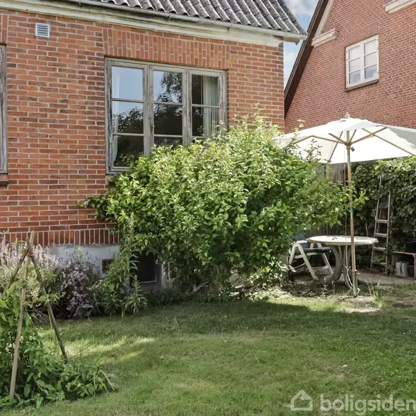 Rødstenshus står i en have med grønne buske og græsplæne. Ved siden af huset er et havebord med en åben parasol. Blomstrende planter vokser langs husets fundament.