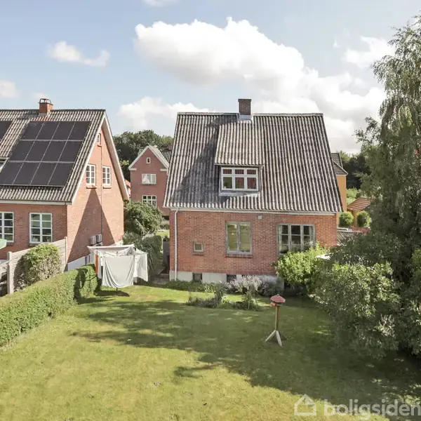 To røde murstenshuse står ved siden af hinanden. Det venstre hus har solpaneler på taget. I forgrunden er en grøn have med hæk og et havemøbel under en parasol.